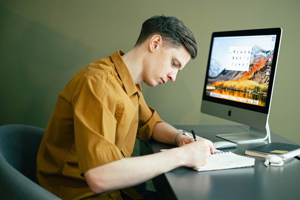 Adult male concentrating on notes with a desktop computer in a home office setting.